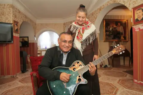 Don Vasyl siedzi z gitarą, obok niego stoi kobieta w chustce.