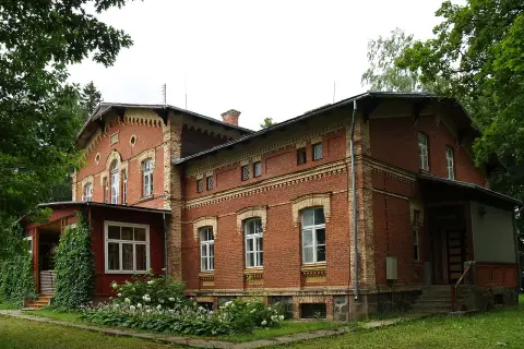 Stary, ceglany maison otoczony zielenią, z werandą i kwitnącymi krzewami.