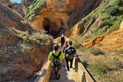 Hikers descend a stone staircase on a scenic trail in Portugal, leading towards a large cave entrance nestled within rugged, sun-drenched cliffs.