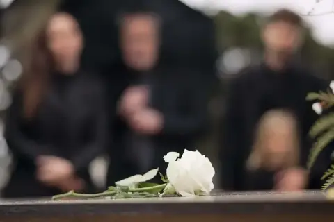 Biała róża na kamiennej płycie, w tle zamazane sylwetki żałobników. Czas organizacji pogrzebu jest trudny.