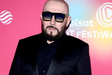 Skawiński w czarnym garniturze i okularach przeciwsłonecznych pozuje na tle różowego banera z logo Polsatu.