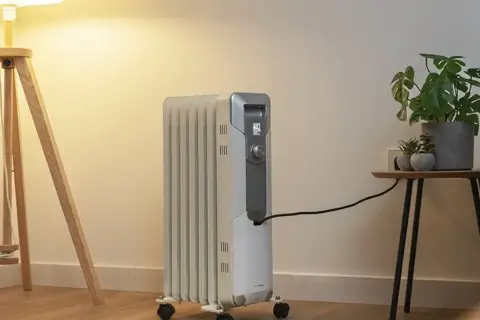 Estufa eléctrica de aceite con ruedas, conectada a la corriente, junto a una planta y una lámpara. Controla el consumo.