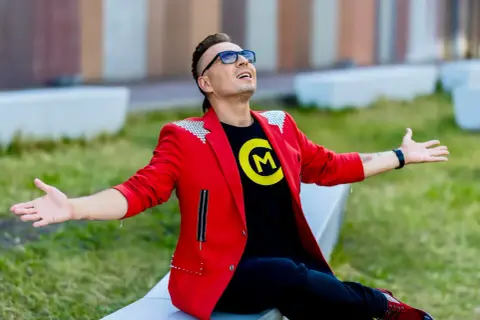 Czadoman w czerwonej marynarce i czarnej koszulce z logo siedzi na ławce z rękami rozłożonymi.