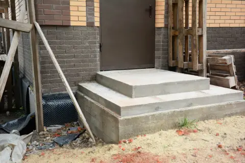 Solidny fundament pod schody z betonu, gotowy na dalsze prace budowlane. Obok widać materiały budowlane i fragment domu.