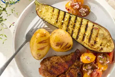 Une demi aubergine au barbecue, des pommes de terre grillées et une salade de tomates sur une assiette blanche.