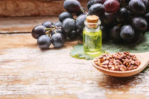 Racimos de uvas oscuras junto a una botella de aceite y una cuchara con semillas, mostrando el origen del extracto de semilla de uva.