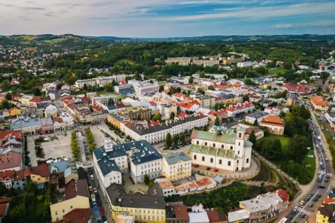 Urokliwe miasto w dolinie ropy, z dominującą białą bazyliką i kolorowymi kamienicami, otoczone zielonymi wzgórzami.