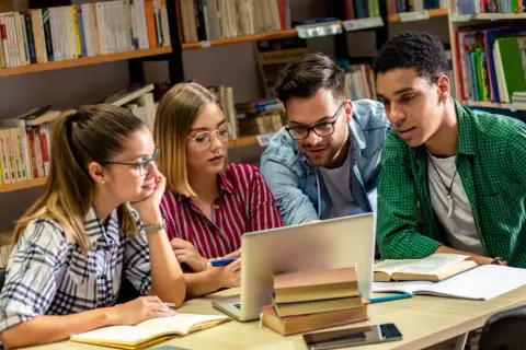Studenci na studiach humanistycznych pochylają się nad laptopem w bibliotece, dyskutując o projekcie.