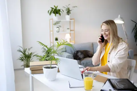 Uśmiechnięta kobieta prowadzi rozmowę telefoniczną, pracując zdalnie okazjonalnie przy laptopie. Na biurku napój i rośliny.