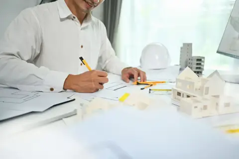 Architekt rysuje plany, obok makieta domu, biały kask i budynki.