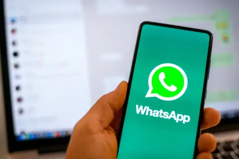 Dłoń trzyma smartfon z logo WhatsApp. W tle widać ekran laptopa z otwartym programem do komunikacji, sugerującym użycie WhatsApp na komputer.