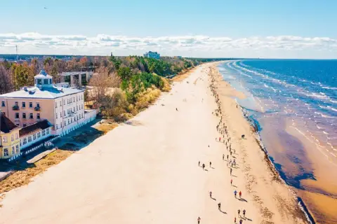 Szeroka, piaszczysta plaża na Łotwie, z budynkami przy brzegu i ludźmi spacerującymi wzdłuż morza.