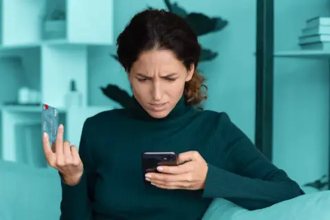 Zmarszczona kobieta trzyma kartę i telefon, zastanawiając się nad bezpieczne zakupy w internecie.