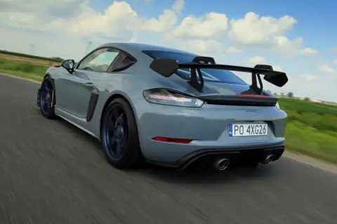 Szary Porsche GT4 RS z dużym skrzydłem pędzi po drodze, zostawiając za sobą rozmyte pola i błękitne niebo.