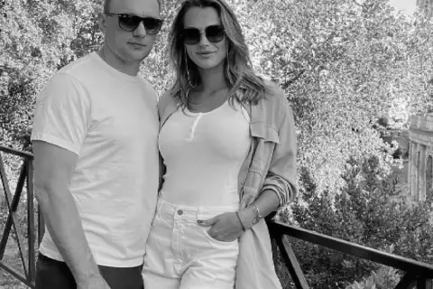 Aryna Sabalenka z partnerem, uśmiechnięci, w okularach przeciwsłonecznych, na tle drzew i architektury.