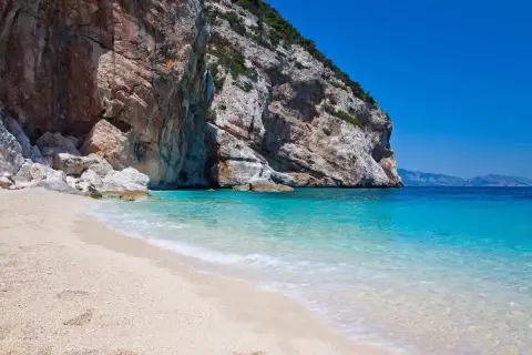 Plage de sable fin bordée par une eau turquoise cristalline, avec une falaise imposante en arrière-plan. Un coin de paradis sur la côte smeralda sardaigne.