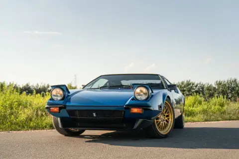 Niebieski De Tomaso Pantera z wyskakującymi reflektorami i złotymi felgami na tle zieleni i błękitnego nieba.