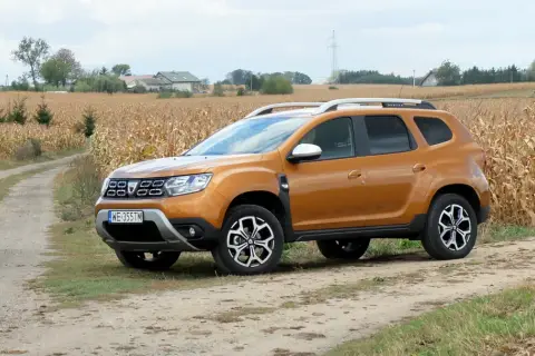 Pomarańczowy Dacia Duster 1.6 benzyna na polnej drodze, w tle pole kukurydzy i domy.