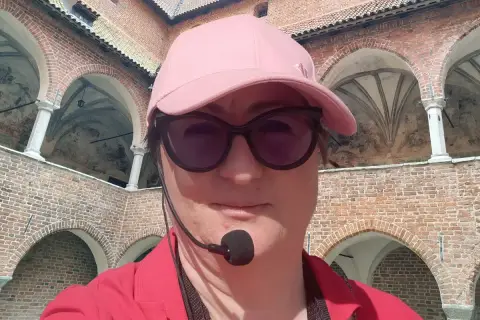 Kobieta w czapce i okularach przeciwsłonecznych zwiedza z przewodnikiem z mikrofonem zamek w Lidzbarku Warmińskim, odkrywając jego atrakcje.