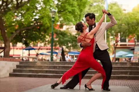 Coppia in abito elegante e vestito rosso esegue una sensuale danza latina all'aperto, tra alberi e gradinate.