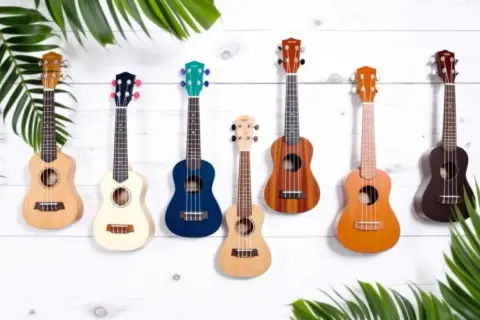 Sept ukulélés de différentes couleurs et tailles, dont un ukulélé de taille soprano, sont accrochés sur un mur en bois blanc, entourés de feuilles vertes.