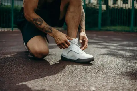 Mężczyzna z tatuażami na ramieniu klęczy, sznurując białe buty sportowe. Może to być przygotowanie do treningu lub ulga po bólu kolan po ćwiczeniach.