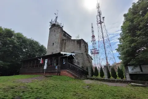 Kamienna wieża i drewniana przybudówka na szczycie góry. W tle wysokie maszty telekomunikacyjne. To popularne miejsce na szlaki turystyczne, jak Chełmiec.