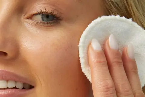 Rostro de mujer rubia con piel luminosa, aplicándose un disco desmaquillante. Su piel luce sin imperfecciones, sin rastro de milium.