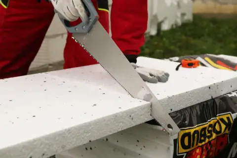 Ręce w rękawiczkach tną styropian piłą ręczną. W tle widać miarkę i materiały budowlane.