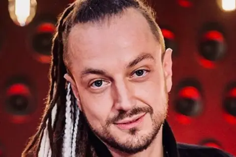 Aleksander Baron z dredami i brodą, na czerwonym tle.