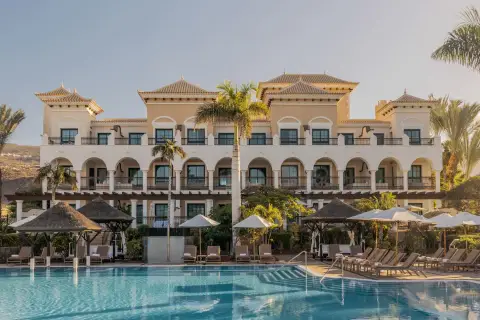 Piscina y edificio del Gran Meliá Palacio de Isora, con palmeras y tumbonas bajo el sol.