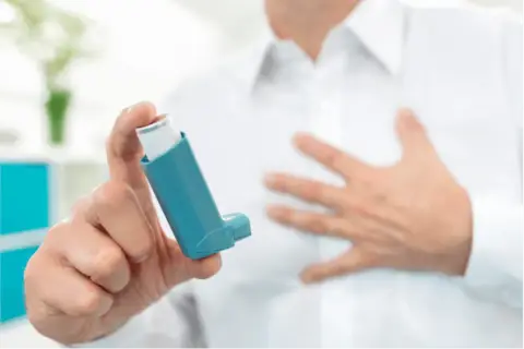 Mann hält Inhalator mit ipratropiumbromid. Seine andere Hand liegt auf der Brust, was auf Atemnot hindeutet.