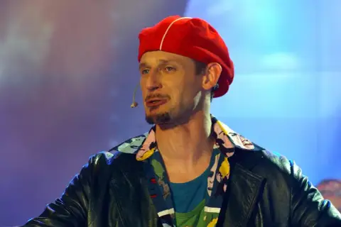 Michał Wójcik w czerwonej beretce i kolorowej koszuli, z mikrofonem przy ustach, na tle niebiesko-fioletowego światła.