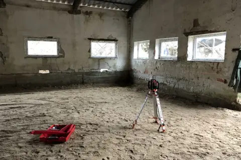Na piaszczystym podłożu stoi niwelator geodezyjny, gotowy do pracy. W tle widać okna i ściany, a na ziemi leży czerwona skrzynka. To przygotowania do wylania posadzki z **gruntu do anhydrytu**.