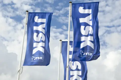 Niebieskie flagi z białym logo JYSK na tle błękitnego nieba z chmurami.