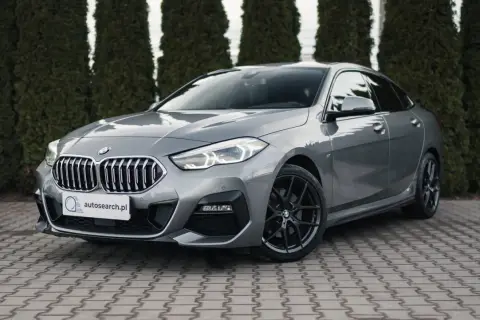 Szary BMW 218i z czarnymi felgami stoi przed żywopłotem.