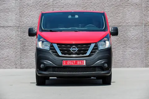Czerwony Nissan NV300 z czarnym zderzakiem i chromowaną atrapą chłodnicy stoi przed betonową ścianą.