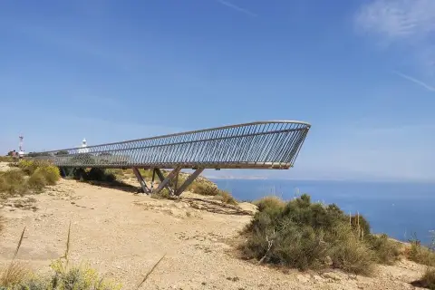 El mirador del faro de Santa Pola se proyecta sobre el mar azul, con vegetación árida y un cielo despejado.