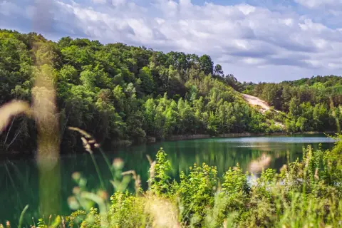 Turkusowe jezioro międzyzdroje otoczone bujną zielenią. W oddali piaszczyste zbocze i błękitne niebo z chmurami.