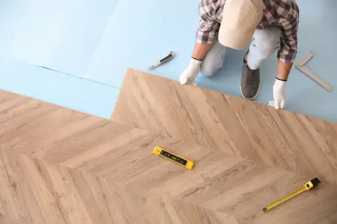 Instalator w białych rękawiczkach układa panele podłogowe w jodełkę. Widać poziomicę i miarkę, co sugeruje, jak kłaść panele.