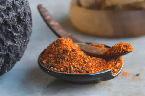 A dark, textured rock sits beside a small bowl filled with a vibrant 7 spice recipe blend. A wooden spoon rests in the bowl, ready to scoop the aromatic mixture.