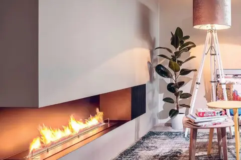 Nowoczesny kominek bio wolnostojący z płonącym ogniem, obok którego stoi lampa i roślina.