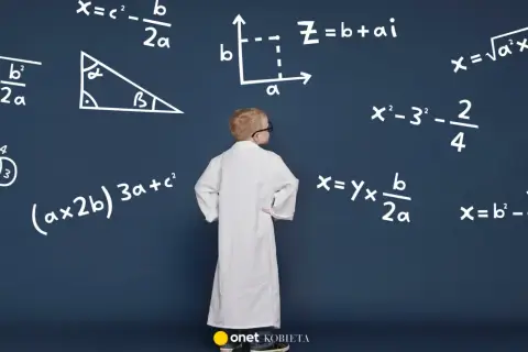 Chłopiec w białym fartuchu stoi przed tablicą z wzorami, w tym wzorem na przyspieszenie.