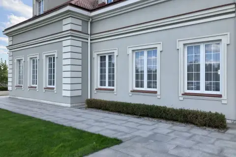 Elegancki budynek z jasnoszarym tynkiem, ozdobiony białymi pilastrami i gzymsami. Okna z białymi ramami i brązowymi parapetami.