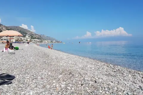Plage de galets à Letojanni, Sicile. Des baigneurs profitent du soleil sous un parasol, tandis que la mer d'un bleu profond s'étend à l'horizon.