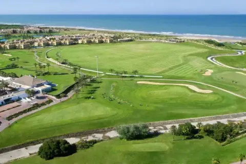 Vista aérea del campo de golf del hotel Barceló Costa Ballena, con el mar azul al fondo y edificios residenciales.