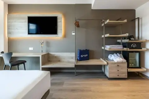 Habitación moderna con TV, escritorio, armario abierto con minibar y caja fuerte. Una bolsa azul con el logo de Senator Hotels descansa sobre un banco.