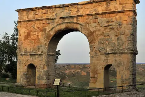 Imponente arco romano de piedra, con dos arcos menores a los lados, se alza sobre un paisaje de colinas verdes.