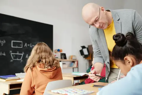 Nauczyciel pomaga uczniom przy biurkach w klasie, na tablicy widać wzory matematyczne.