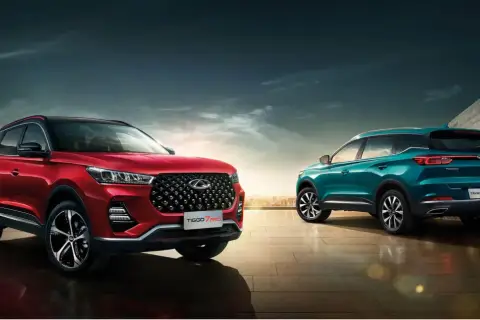 Dwa stylowe chińskie samochody, czerwony i turkusowy SUV, prezentują się na tle miejskiego krajobrazu i nowoczesnej architektury.